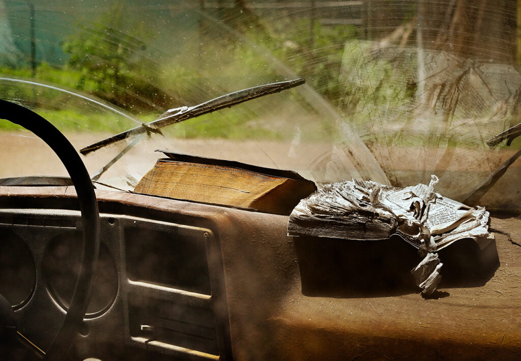 A color image of the inside dashboard in tones of brown. A portion of the steering wheel is seen and the wipers are stopped mdi-swipe. A thick bible with the spine toward the windshield is center left on the dash. An open, very worn bible with curled pages is center right on the dash