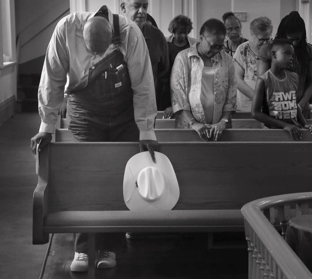 Black and white image of inside a church. First pew is empty. A black man in overalls with one hook undone and a cowboy hat in his hand, head bowed, is in second pew. Several other people are seen, including an older black man, three older black women, a white woma, an younger black woman, and an adolescent black boy.