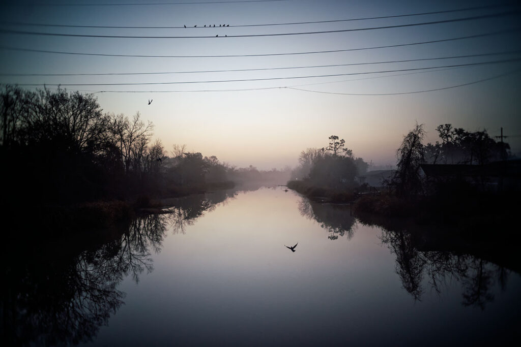 moody image of a still river with shrubs and trees on each bank. There are power lines across the top with birds on them. A bird is seen flying just above the river.